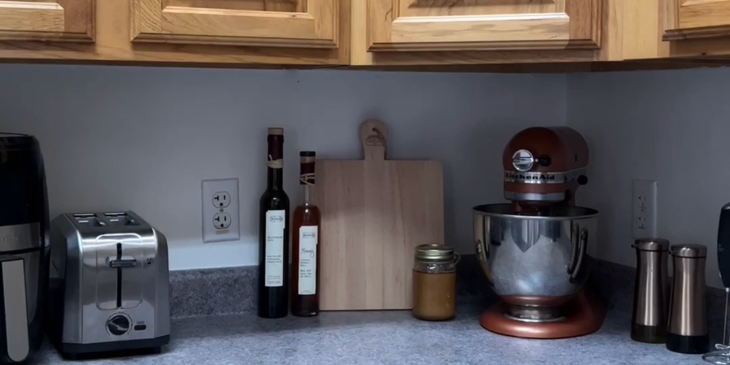 A modern kitchen counter featuring a silver toaster, a KitchenAid mixer, oil and vinegar bottles, a cutting board, and a jar of honey.