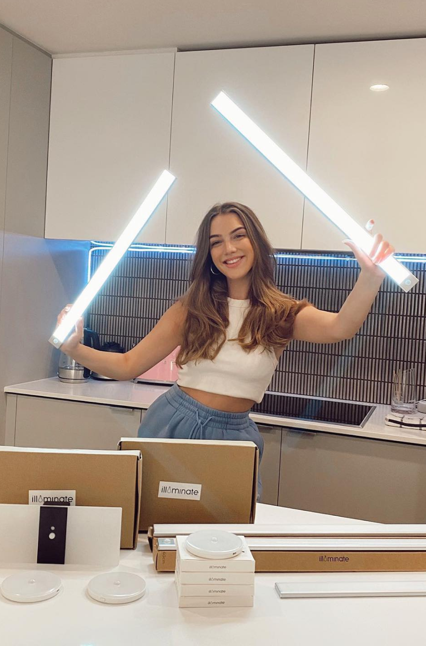 A woman poses in a modern kitchen, holding glowing light tubes, with illuminated packaging of lighting products scattered on the counter.