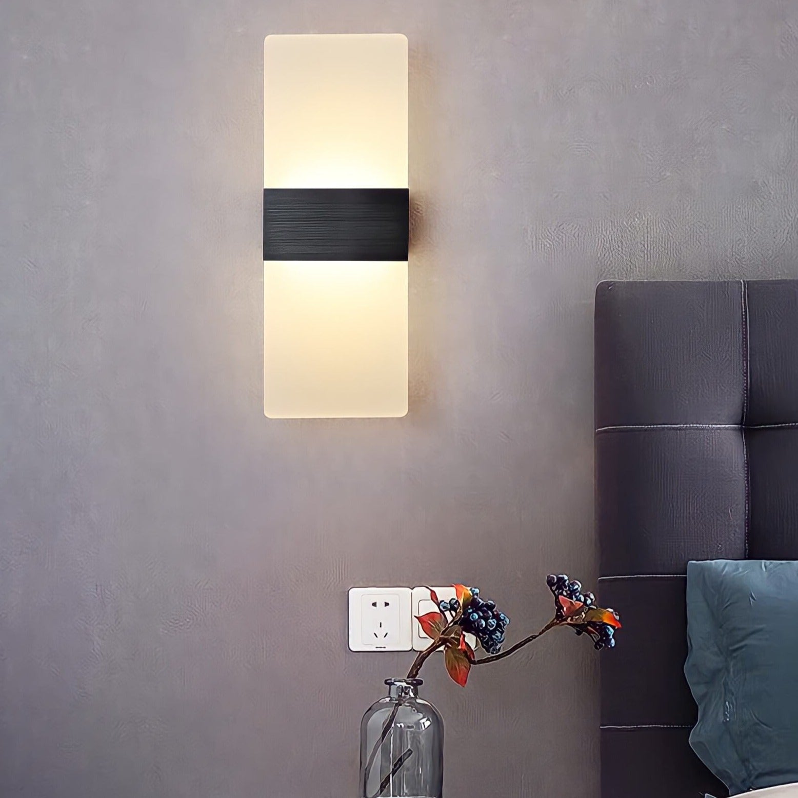 A modern wall lamp illuminates a gray wall above a bedside table with a glass vase and a book, creating a cozy bedroom ambiance.
